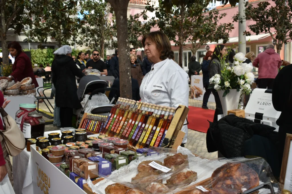 Banca com frascos de compotas, doces embalados, licores e pão artesanal, atendida por produtora local.