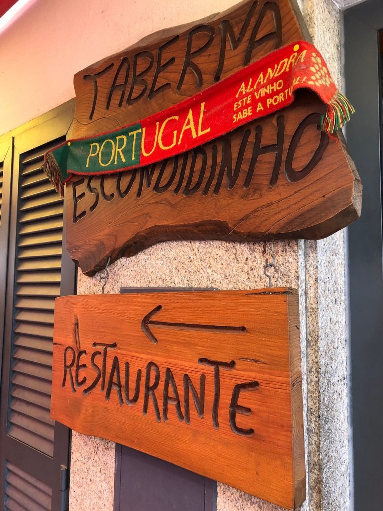 Placas de madeira fixadas numa parede de pedra, indicando “Taberna Escondidinho” e “Restaurante”, com um cachecol nas cores de Portugal sobre a placa superior.