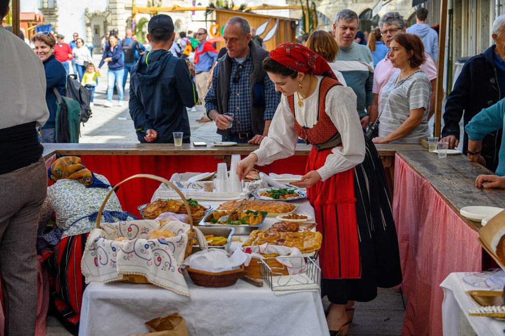 Mesa com pratos de pataniscas, doces e pão, organizada por mulher em traje tradicional vermelho.