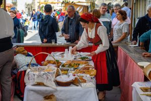 Mesa com pratos de pataniscas, doces e pão, organizada por mulher em traje tradicional vermelho.