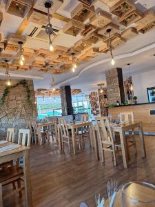 Interior do restaurante com mesas de madeira, paredes em pedra, plantas e teto decorado com caixas de madeira e lâmpadas suspensas.