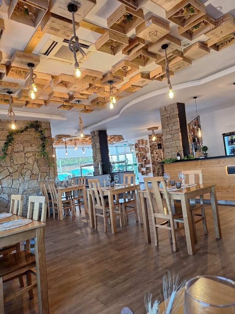 Interior do restaurante com mesas de madeira, paredes em pedra, plantas e teto decorado com caixas de madeira e lâmpadas suspensas.