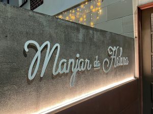 Placa de parede com o nome “Manjar da Helena” em letra cursiva, iluminada por luzes quentes suspensas.