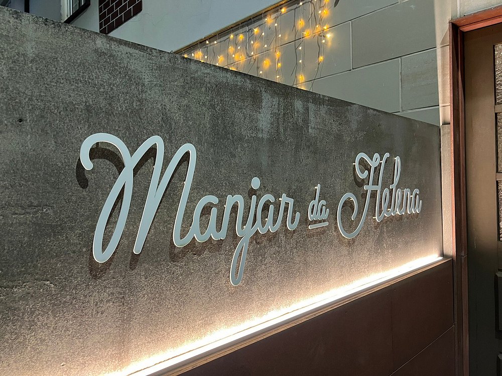 Placa de parede com o nome “Manjar da Helena” em letra cursiva, iluminada por luzes quentes suspensas.