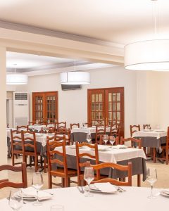 Sala de refeições com mesas cobertas por toalhas brancas e cinzentas, com copos, pratos e talheres dispostos para receber clientes.