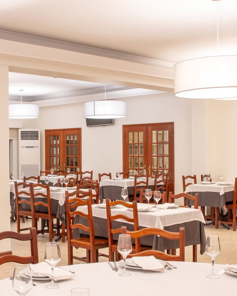 Sala de refeições com mesas cobertas por toalhas brancas e cinzentas, com copos, pratos e talheres dispostos para receber clientes.