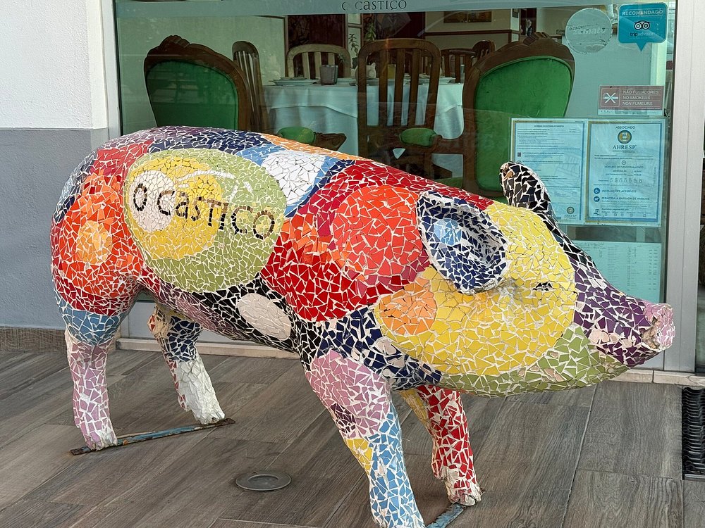 Escultura de porco revestida a mosaicos coloridos colocada à entrada do restaurante O Castiço, com interior visível ao fundo.