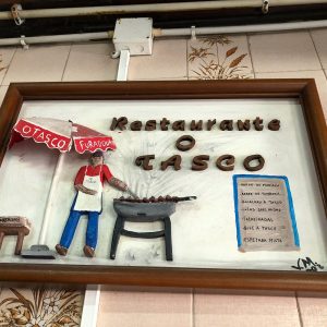 Placa decorativa com figura a grelhar alimentos e lista de pratos do Restaurante O Tasco.