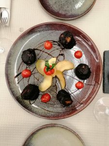 Prato redondo com peças de carne, fruta laminada no centro, tomate e fios de molho escuro.