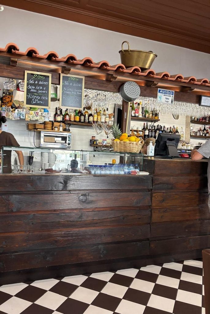 Interior com balcão de madeira, garrafas expostas, fruta fresca e quadros de menu escritos a giz.