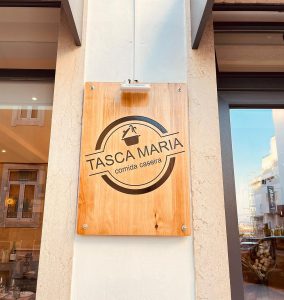 Placa de madeira com o nome “Tasca Maria” e o texto “comida caseira” fixada numa parede exterior.
