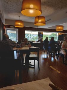 Sala de refeições com várias mesas ocupadas por clientes, iluminação suspensa e janelas amplas.