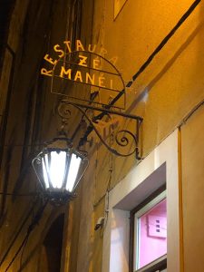 Placa metálica com o nome do restaurante, iluminada por um candeeiro de rua numa parede amarela.
