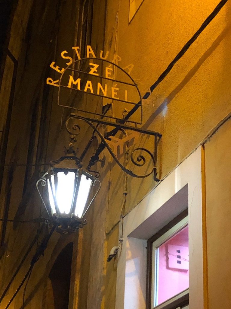 Placa metálica com o nome do restaurante, iluminada por um candeeiro de rua numa parede amarela.