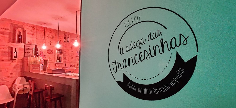 Balcão com bancos altos, garrafas expostas, iluminação pendente e parede turquesa com o logótipo “A Adega das Francesinhas”.