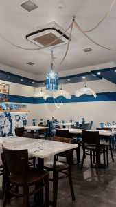 Sala com mesas e cadeiras de madeira, paredes e teto com faixas azuis, candeeiros decorativos, cordas suspensas e balcão com elementos artísticos.