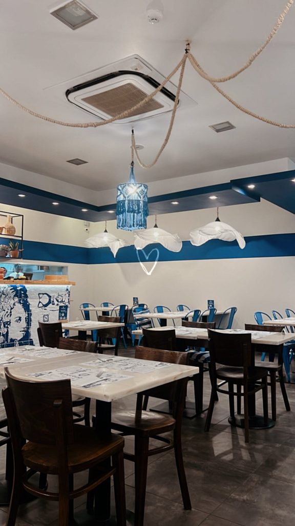 Sala com mesas e cadeiras de madeira, paredes e teto com faixas azuis, candeeiros decorativos, cordas suspensas e balcão com elementos artísticos.