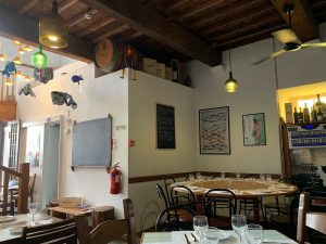 Interior de restaurante com mesas montadas, luzes coloridas, vigas de madeira, garrafas de vinho expostas e decoração com peixes suspensos.