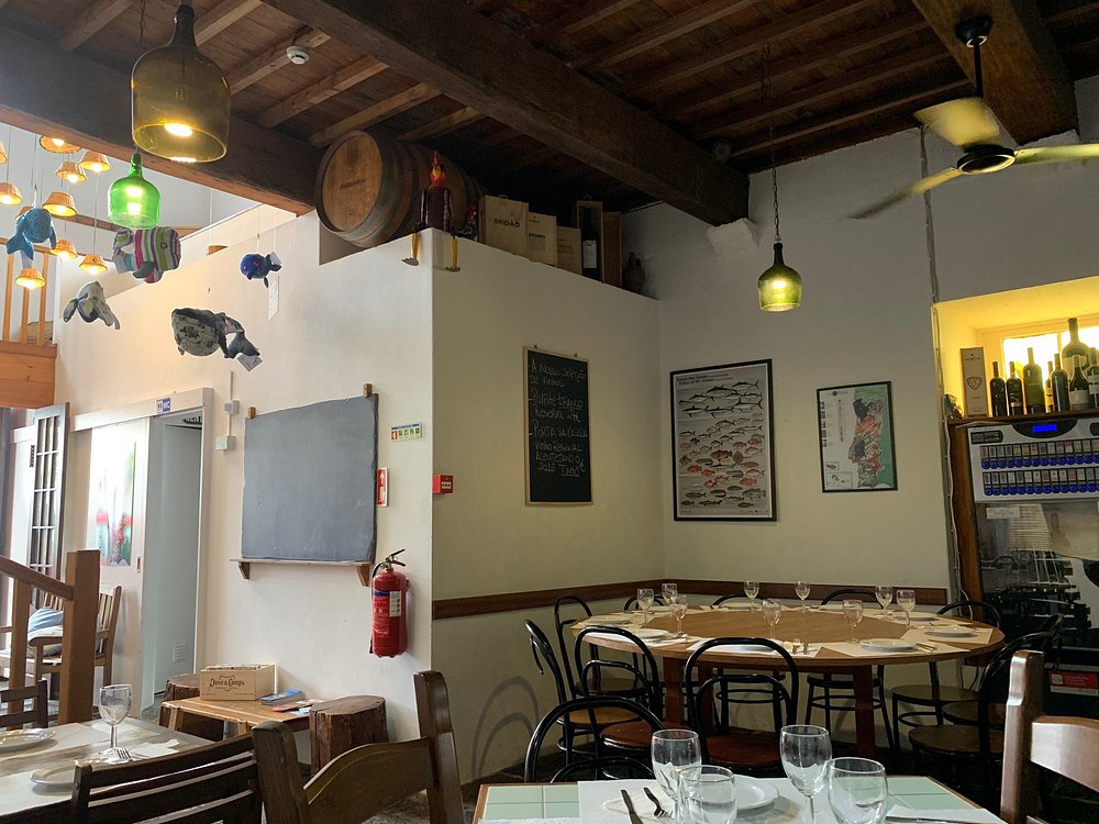 Interior de restaurante com mesas montadas, luzes coloridas, vigas de madeira, garrafas de vinho expostas e decoração com peixes suspensos.