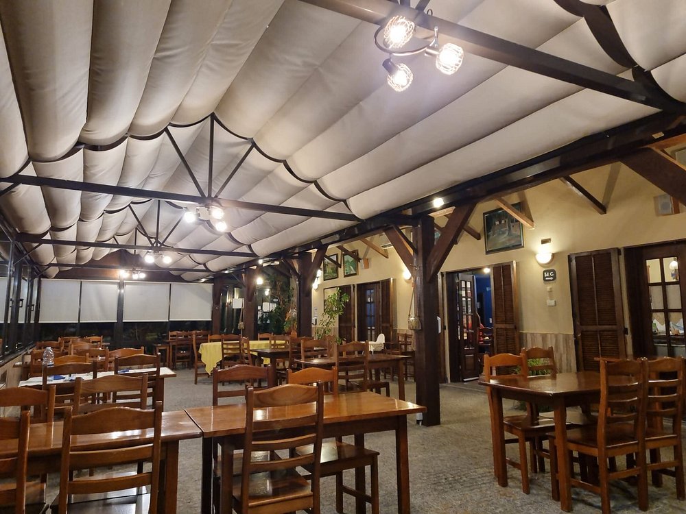 Sala com mesas de madeira, cadeiras, teto com tecido branco, quadros e plantas.