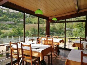 Sala com mesas de madeira, toalhas brancas e janelas grandes com vista para rio e colinas verdes.