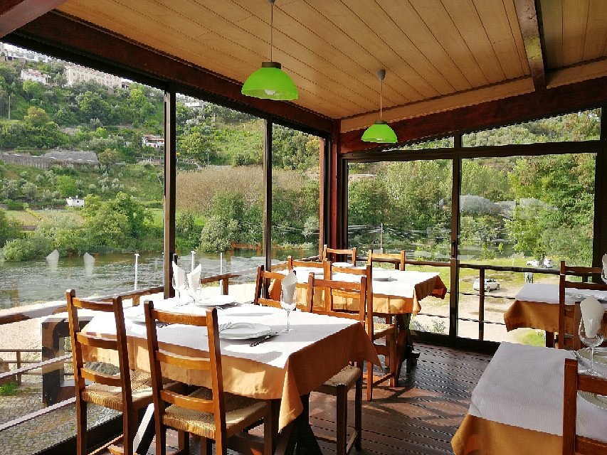 Sala com mesas de madeira, toalhas brancas e janelas grandes com vista para rio e colinas verdes.