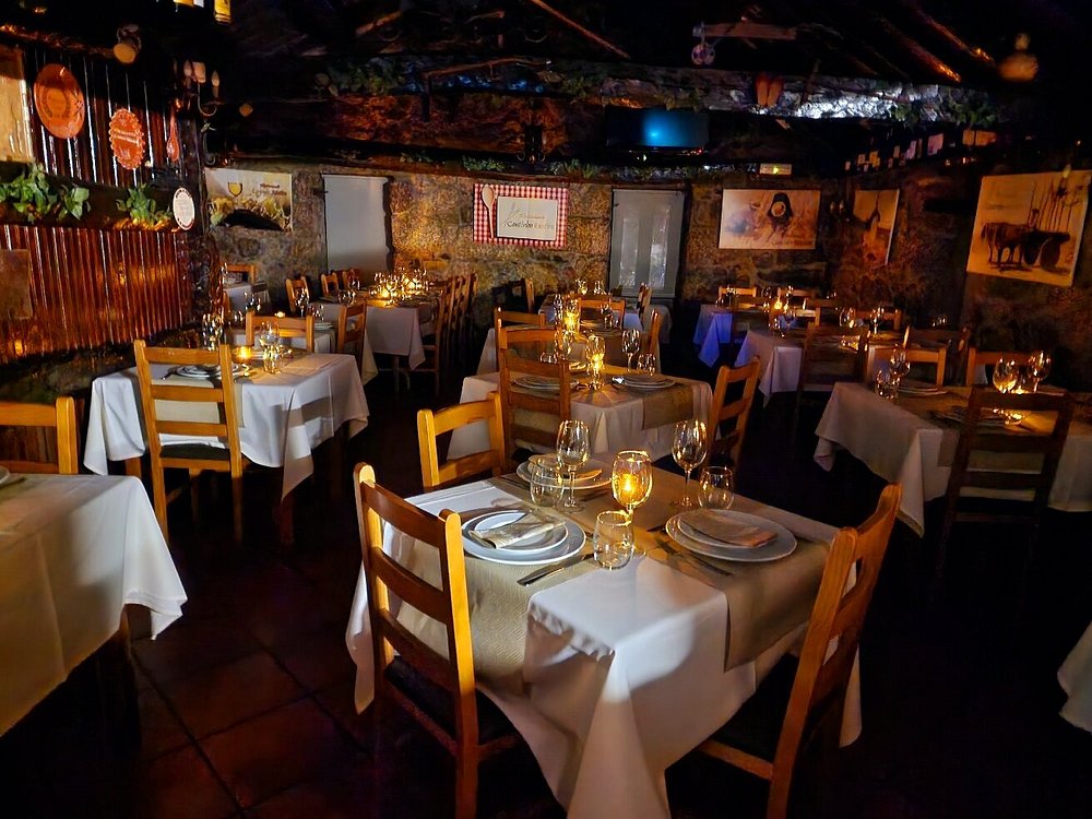 Sala com mesas de madeira cobertas por toalhas claras, velas acesas, paredes de pedra com quadros e plantas, e teto com vigas de madeira.