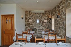 Sala de restaurante com paredes de pedra, mesas com toalhas brancas e corredores com toalhas axadrezadas, cadeiras de madeira e iluminação quente.
