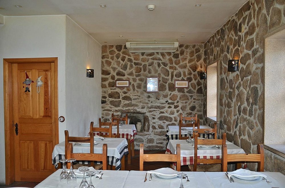 Sala de restaurante com paredes de pedra, mesas com toalhas brancas e corredores com toalhas axadrezadas, cadeiras de madeira e iluminação quente.