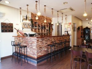 Interior de restaurante com balcão em pedra, iluminação com lâmpadas suspensas, garrafas expostas e mesas de madeira.