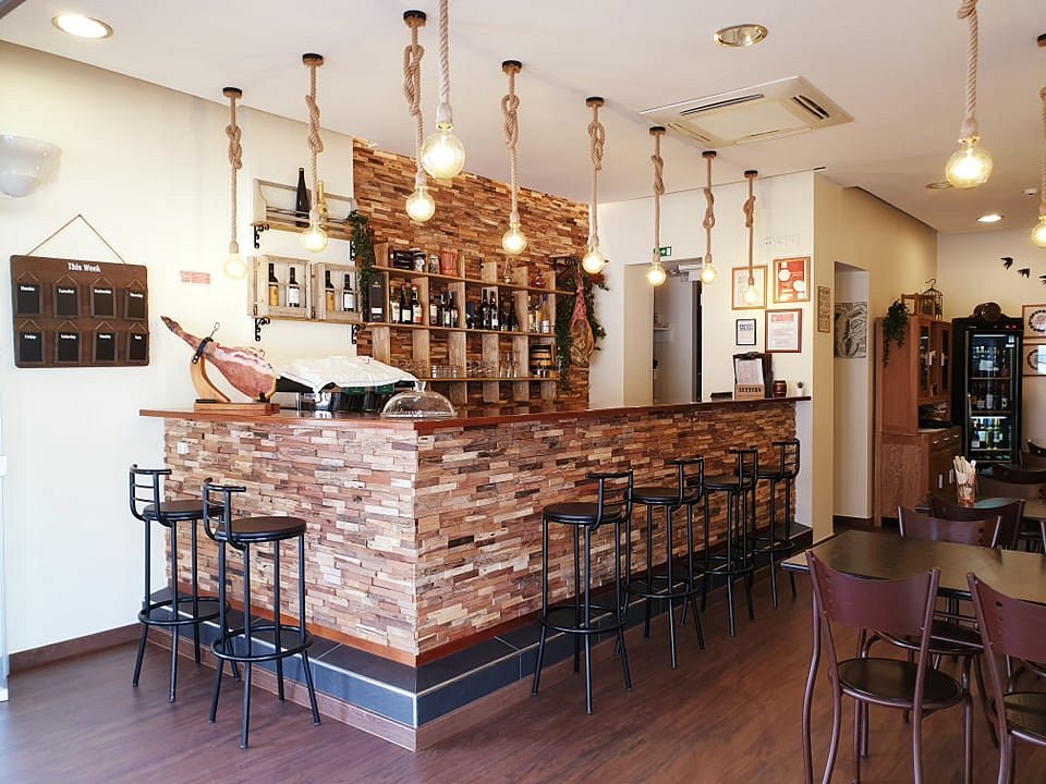 Interior de restaurante com balcão em pedra, iluminação com lâmpadas suspensas, garrafas expostas e mesas de madeira.