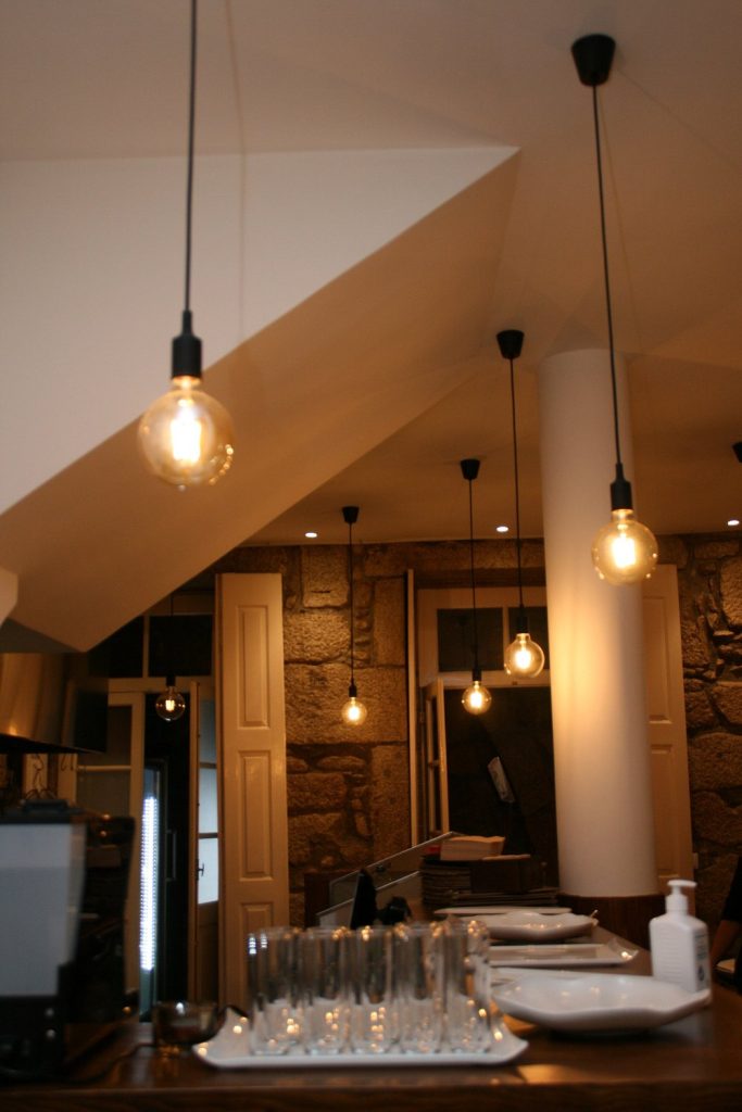 Sala com lâmpadas pendentes de luz quente, teto branco com coluna central, balcão com copos, pratos e desinfetante; ao fundo paredes de pedra, portas abertas e mais iluminação.