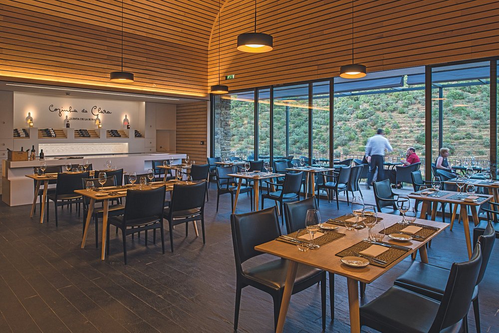 Sala de restaurante com mesas montadas, cadeiras escuras e janelas grandes com vista para vinhas em socalcos.