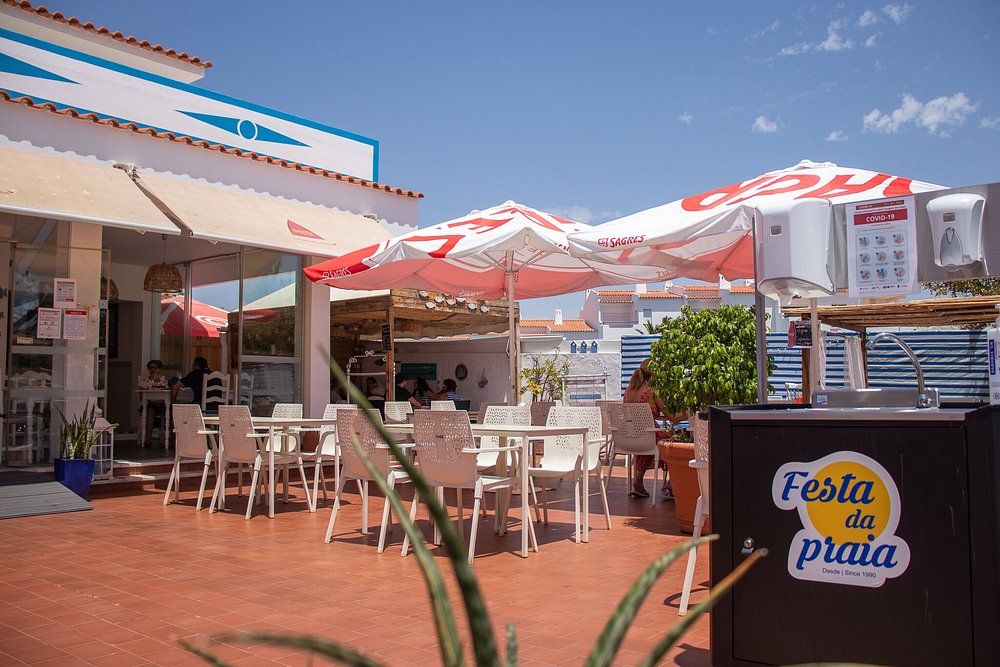 Esplanada com mesas brancas, chapéus de sol Sagres e clientes sentados num ambiente ensolarado.