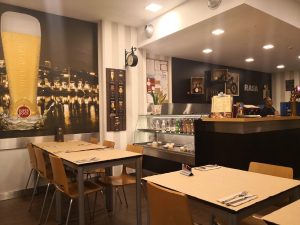 Sala com mesas de madeira e individuais bege, cartaz de cerveja na parede, balcão com vitrina de bebidas e sobremesas, prateleiras com garrafas e decoração, palavra “RASA” na parede e funcionário atrás do balcão.