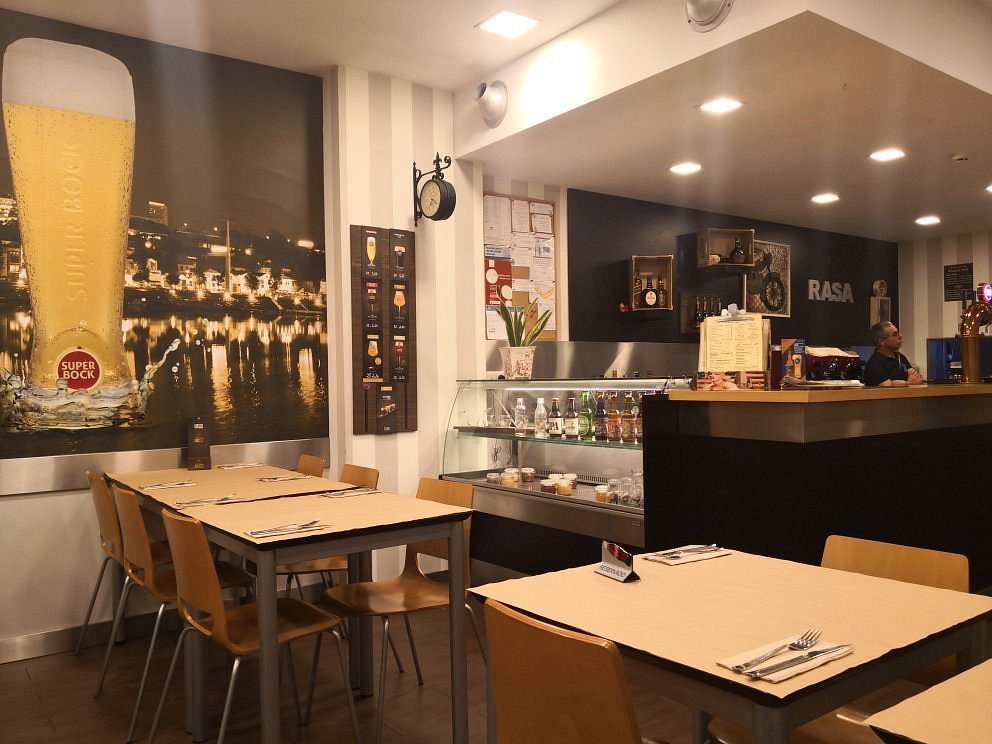 Sala com mesas de madeira e individuais bege, cartaz de cerveja na parede, balcão com vitrina de bebidas e sobremesas, prateleiras com garrafas e decoração, palavra “RASA” na parede e funcionário atrás do balcão.