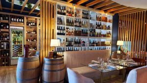 Sala com prateleiras de vinhos, barris de madeira, mesa com tampo de vidro, pratos, copos e iluminação quente.