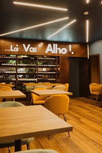 Sala de restaurante com mesas de madeira, cadeiras amarelas e verdes, teto com luzes lineares e parede com letreiro iluminado “…Lá Vai’ Alho!” sobre garrafeira com garrafas e plantas.