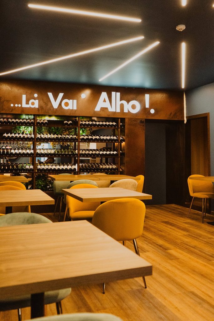 Sala de restaurante com mesas de madeira, cadeiras amarelas e verdes, teto com luzes lineares e parede com letreiro iluminado “…Lá Vai’ Alho!” sobre garrafeira com garrafas e plantas.