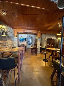 Interior de restaurante com teto em madeira, paredes em pedra, mesas e cadeiras rústicas, iluminação quente e arco em pedra ao fundo.