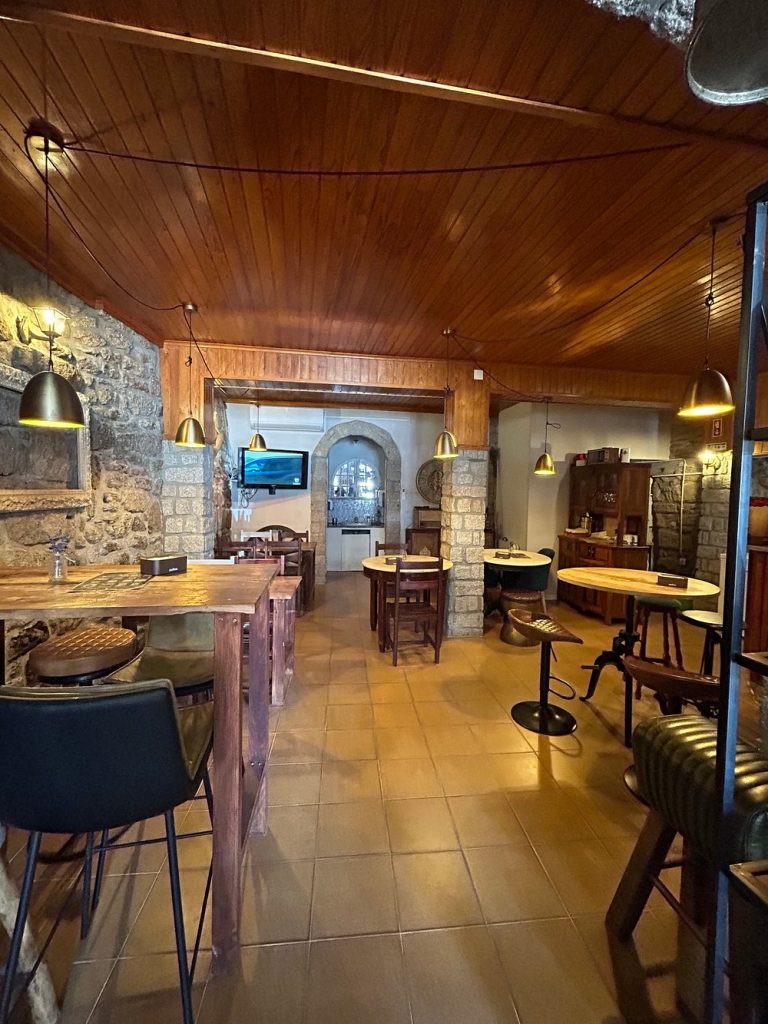Interior de restaurante com teto em madeira, paredes em pedra, mesas e cadeiras rústicas, iluminação quente e arco em pedra ao fundo.