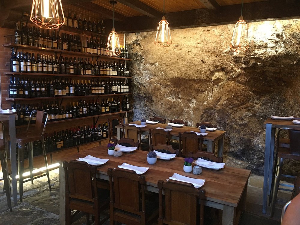 Sala com mesas de madeira, garrafeira com garrafas alinhadas e parede de pedra iluminada por candeeiros metálicos.