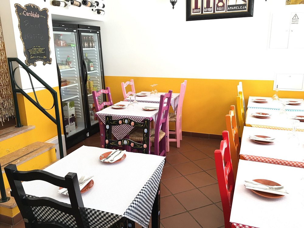 Sala pequena com mesas cobertas por toalhas aos quadrados de várias cores, cadeiras pintadas em tons vivos, frigorífico com porta de vidro, garrafas expostas e quadro com “Cardápio” na parede.