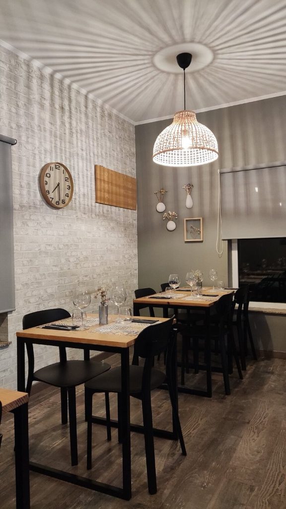 Sala com mesas de madeira, cadeiras pretas, parede em tijolo cinzento e candeeiro decorativo.