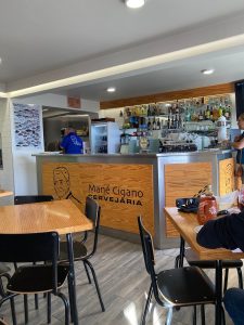 Interior da Cervejaria Mané Cigano com balcão em madeira e metal, garrafeira, máquina de café, mesas de madeira e clientes.