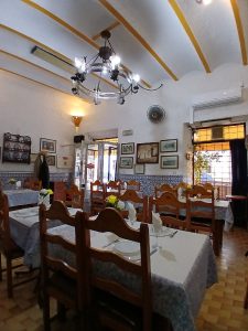Sala com mesas de madeira, toalhas estampadas, copos, talheres, quadros e azulejos azuis e brancos.