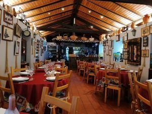 Sala com mesas cobertas por toalhas vermelhas, paredes com objetos decorativos e bar com garrafas ao fundo.