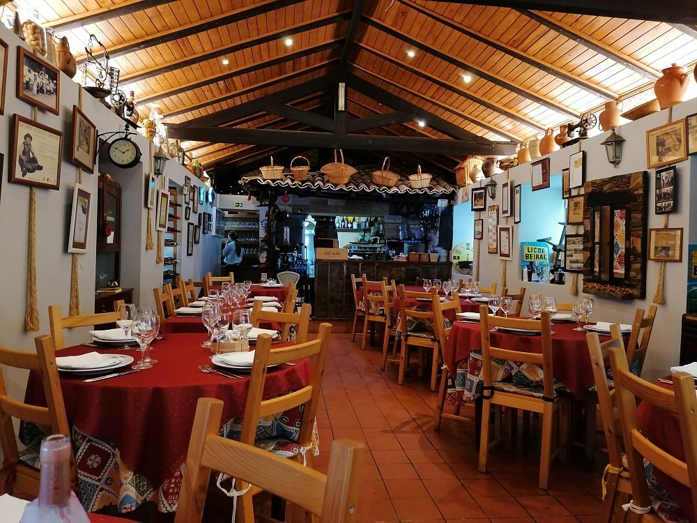 Sala com mesas cobertas por toalhas vermelhas, paredes com objetos decorativos e bar com garrafas ao fundo.