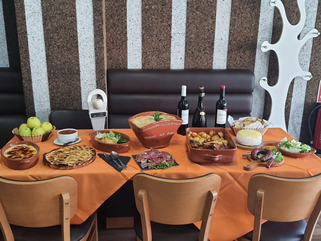 Mesa com vários pratos tradicionais, incluindo arroz no forno, carnes assadas, enchidos, sobremesas, pão, queijo, maçãs e garrafas de vinho.
