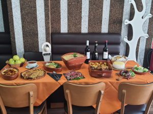 Mesa com vários pratos tradicionais, incluindo arroz no forno, carnes assadas, enchidos, sobremesas, pão, queijo, maçãs e garrafas de vinho.